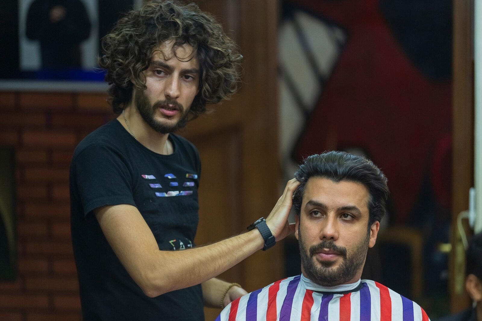 Un barbero arregla el pelo de un cliente con capa a rayas, en una barbería